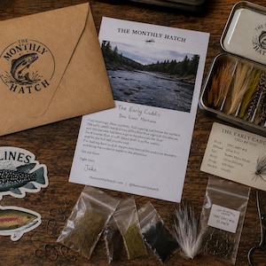May include: A fly fishing kit including a brown envelope with The Monthly Hatch logo, an informational card about The Early Caddis, a metal tin containing assorted feathers, two fish stickers, small bags of dubbing, hooks, and scissors.