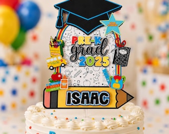 May include: A cake topper featuring a black graduation cap, a rainbow arch, and school-themed icons including a yellow bus, crayons, books, and a backpack. The text reads PRE-K grad 2025 and ISAAC. It is displayed on a white frosted cake with letters and numbers.