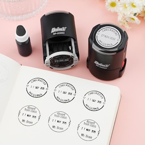 May include: A black self-inking stamp with the text "CHECKED BY" and "MS GUINA" is shown next to a notebook with stamped impressions. A small ink bottle is also present. The stamps are for office or classroom use.
