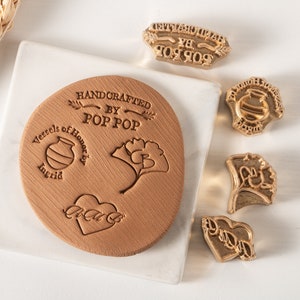 May include: A set of five gold-colored metal stamps with various designs. The stamps are used to create impressions on leather or other materials. The designs include a heart, a ginkgo leaf, a pot, and two different text stamps. The text stamps read "Handcrafted by Pop Pop" and "Vessels of Honor by Ingrid".