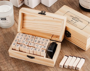 May include: A wooden alphabet and number stamp set with a black ink pad. The set includes individual letter and number stamps, stored in a wooden box. The box is open, revealing the stamps. Additional stamps are visible outside the box.