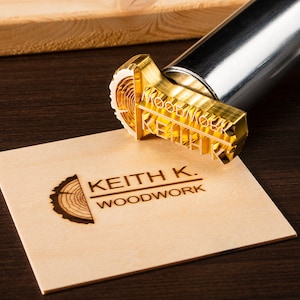 May include: A gold-colored metal branding iron with the text "KEITH K. WOODWORK" engraved on it. The branding iron is resting on a piece of wood with the same logo burned into it.