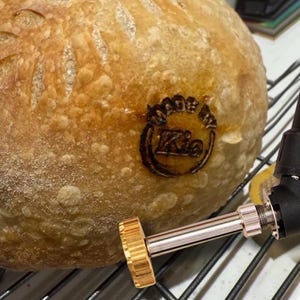 May include: A loaf of artisan bread with a golden-brown crust. The bread has a decorative pattern on top and a branding mark that reads "Made by Kie". A metal tool is in the foreground.