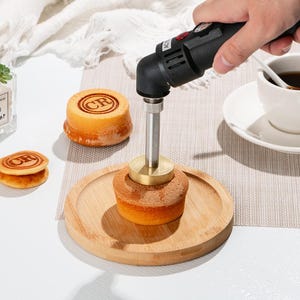 Puede incluir: Primer plano de un hierro de marcar de mano que se utiliza para estampar un diseño en un pequeño pastel. El pastel está en un plato de madera. El hierro de marcar tiene una varilla de metal con una punta de latón y un mango negro con un botón rojo. El pastel es redondo y tiene un color marrón claro.