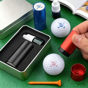 May include: A golf gift set featuring a black and red golf ball marker, two white golf balls with the text "JAMES GOLF", a blue marker holder, and an orange tee. The set is presented on a green surface.