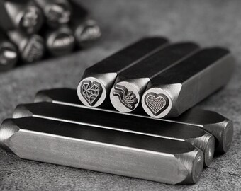 May include: Close-up of metal stamps with various heart and floral designs. The stamps are arranged in a row, with the detailed designs visible on the stamp heads. The tools have a rectangular shape and are made of a silver-colored metal.