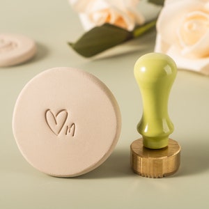 May include: A light pink clay stamp with a heart and the letter "M" imprinted on it. A green wooden handle with a brass base is next to the stamp.