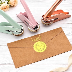 May include: Three wax seal stamps in pastel green, pink, and rose gold are displayed with a brown envelope. The envelope features a gold foil seal with the letter "T" inside a laurel wreath design. A ribbon and flowers are in the background.