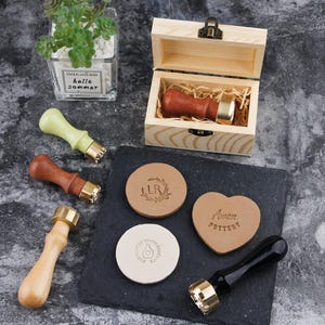 May include: A collection of wax seal stamps with wooden handles and brass stamp heads. The stamps are in a wooden box and next to stamped leather pieces. The leather pieces have designs and text. A small plant in a glass container is in the background.