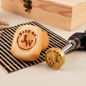 May include: A close-up of a wooden branding iron with the text "MADE BY JW" on the top. The branding iron is on a bamboo mat next to a round bread roll with the same branding.