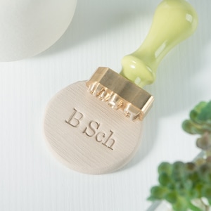 May include: A brass stamp with a wooden handle, used to create an imprint of the text "B.Sch" on a light brown surface.