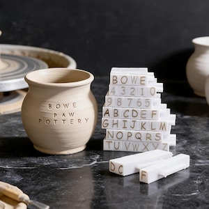 May include: A beige pottery vase with the text "BOWE PAW POTTERY" is next to a stack of white letter and number tiles. Two white letter stamps are in front of the tiles. The pottery is handmade.