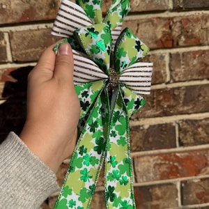May include: A festive St. Patrick's Day bow featuring green and white ribbons with shamrock patterns and gold accents. The bow is held against a brick wall, showcasing its vibrant colors and design.