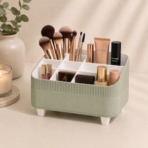 May include: A sage green rectangular makeup organizer with a ribbed exterior and four small white feet. The interior features multiple white dividers holding various makeup brushes, tubes of lipstick, a foundation bottle, and compact cases.