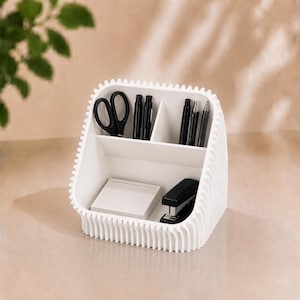 Op de afbeelding: Een witte bureau-organizer met een geribbeld exterieurontwerp en meerdere vakken. De organizer bevat een zwarte schaar, vier zwarte pennen, een stapel wit rechthoekig notitiepapier en een zwarte nietmachine in de verschillende secties.