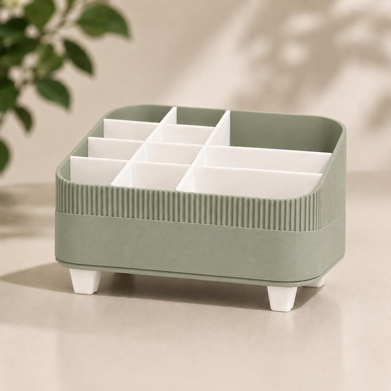 May include: A sage green desk organizer featuring a rectangular shape with rounded corners and a ribbed exterior texture. The interior is divided into multiple white compartments of varying sizes for storage, supported by four small white feet.