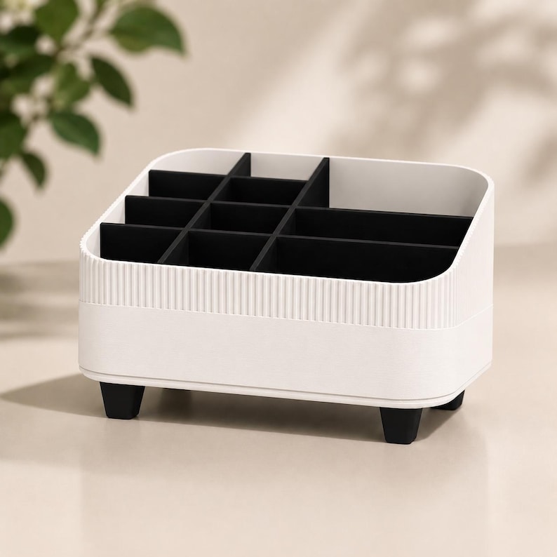 May include: A white desk organizer with a ribbed exterior texture and four black feet. The interior features a black grid of dividers creating multiple compartments of varying sizes for storage, with a sloped top edge design for easy access.