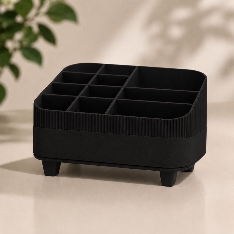 May include: A matte black desktop organizer featuring multiple compartments of varying sizes. The exterior has a vertical ribbed texture along the upper section and a smooth base. The unit is supported by four small, sturdy feet at the corners.