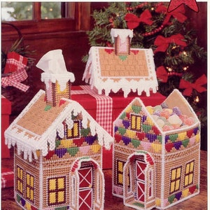 May include: A collection of handcrafted, colorful, plastic canvas Christmas cottages. The cottages feature brown, tan, and white roofs, with colorful walls and red doors. The image includes the text "Enchanted Cottage" and a red star that says "Christmas".