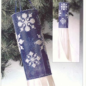 May include: A blue and white plastic canvas windsock with a snowflake design. The windsock is hanging from a tree branch and has white ribbons at the bottom.