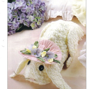 May include: A white crocheted floppy ear bunny wearing a pink and green straw hat with purple and yellow flowers. The bunny is lying on a white surface with a bouquet of purple flowers in the background.