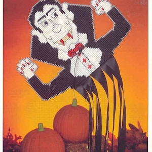 May include: Count Scarey plastic canvas pattern featuring a vampire with fangs and a cape. The pattern is surrounded by a yellow and orange background with two pumpkins in the foreground.