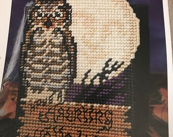 May include: A cross-stitch pattern featuring a white ghost with a black cat in front of a purple and black background. The text "Happy Halloween" is stitched in brown on a tan background.