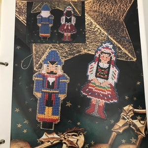 May include: A page from a craft book titled "NOSTALGIC ORNAMENTS" featuring two plastic canvas Christmas ornaments. One is a nutcracker in blue, red, and yellow. The other is a girl in a red skirt and white top. The background is dark green with gold stars.