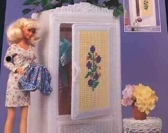May include: A white wicker wardrobe with a yellow and floral cross-stitch door panel. The wardrobe is decorated with faux greenery and is next to a doll holding a hanger with clothes. A small wicker table holds a pot of flowers. The text "Wicker Wardrobe" is at the top.