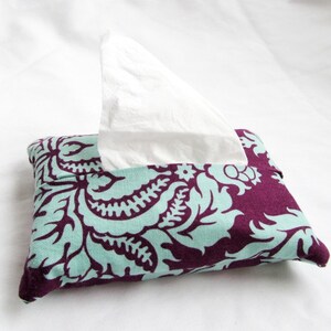 Könnte beinhalten: Ein rechteckiger Taschentuchbox-Bezug mit einem teal- und lila-farbenen Blumenmuster. Weiße Taschentücher ragen aus der oberen Öffnung heraus. Der Stoff hat ein Damast-Design. Der Taschentuchbox-Bezug liegt auf einer weißen Oberfläche.