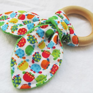May include: A baby teething ring with a wooden ring and fabric bunny ears. The fabric features a colorful turtle pattern on a white background, with green trim. The wooden ring is smooth and light-colored.