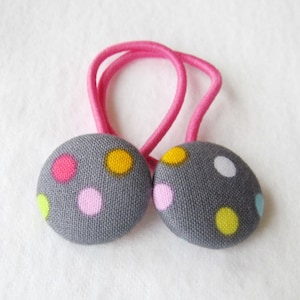 May include: Two gray fabric-covered hair ties with colorful polka dots. Each tie features a round, button-like design with pink, yellow, and other colored dots. The elastic band is a bright pink color.