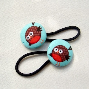 May include: Two hair ties with round, light blue fabric-covered buttons. Each button features a cartoon robin with a brown body, red breast, and white eyes. The hair ties are black and are set against a white background.
