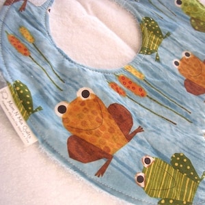 Könnte beinhalten: Baby-Lätzchen mit blauem Hintergrund mit Cartoon-Fröschen, Schilf und Wasserwellen. Die Frösche sind braun und grün mit großen Augen. Das Lätzchen hat eine weiße Rückseite und ein Etikett mit der Aufschrift "Mom N Mia Quilts."