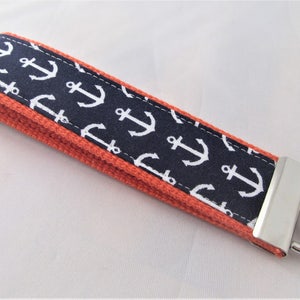 May include: A navy blue and white anchor pattern key fob with a bright orange border and a silver-colored metal clasp and key ring. The key fob is a nautical-themed accessory.