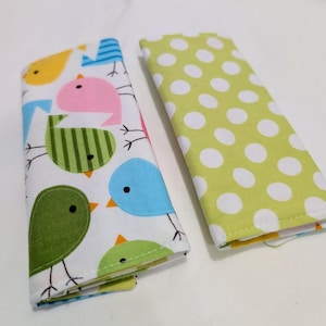 May include: Two fabric cases. One is white with colorful bird illustrations in yellow, pink, green, and blue. The other is light green with white polka dots. Both cases are rectangular and folded.