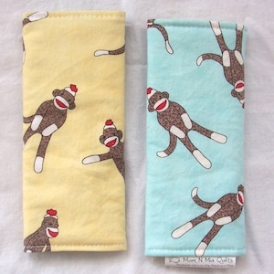 May include: Two rectangular fabric cases, one yellow and one light blue, each patterned with brown sock monkey figures wearing red hats. The cases are likely for eyeglasses or similar items. The light blue case has a small tag that reads "Mom N Mia Quilts".