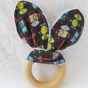 May include: A baby teether with a wooden ring and fabric ears. The fabric is black with a pattern of colorful robots and yellow circles. The ears have a light blue fluffy trim.
