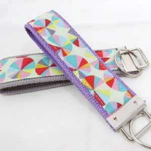 May include: Two key fobs with a colorful geometric pattern. The fabric features a repeating design of multicolored semi-circles on a light green background. The edges are trimmed with a contrasting purple and gray fabric, and each has a silver metal key ring.