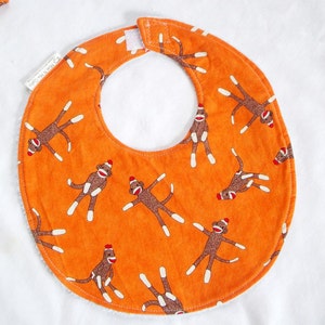 May include: Orange baby bib with a pattern of sock monkeys in various poses. The bib has a white terry cloth backing and a Velcro closure at the neck. The bib is approximately 8 inches wide.