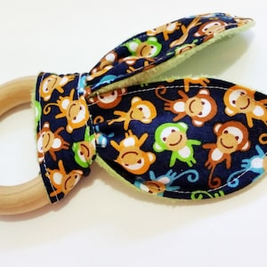 Op de afbeelding: Een houten bijtring voor baby's met stoffen oren. De stof is marineblauw met een patroon van oranje, groene en witte apen. De bijtring is ontworpen voor baby's en jonge kinderen.