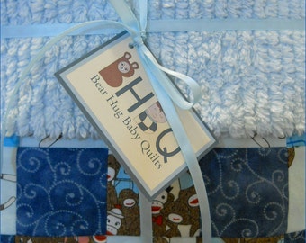 Puede incluir: Una colcha para bebé con una parte superior de felpa azul y una parte inferior de retazos con diseños de monos calcetín. La colcha está atada con una cinta azul claro y una etiqueta que dice "Bear Hug Baby Quilts".