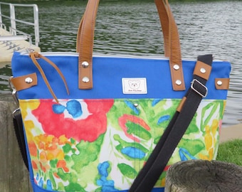 May include: Blue tote bag with a floral pattern in red, green, and yellow. It features brown leather handles and accents, a black shoulder strap, and a zip closure. A small tag with a bear logo is attached.