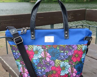 May include: A blue tote bag with black leather handles and a black adjustable shoulder strap. The bag has a colourful floral pattern on the front, a black base, and a label that reads "Bear The Dogs".