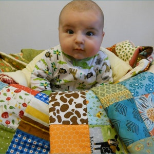 May include: A baby wearing a white and green animal-print onesie sits on a colorful patchwork quilt. The quilt features various patterns, including floral, mushroom, and geometric designs, in shades of blue, red, yellow, and brown.