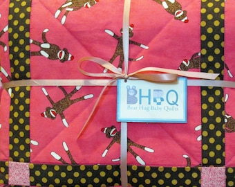 Puede incluir: Una colcha rosa para bebé con un patrón repetido de monos calcetín. La colcha está bordeada con una tela de lunares marrones y dorados y atada con una cinta rosa. Una etiqueta dice "Bear Hug Baby Quilts".