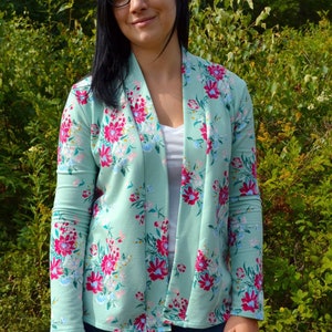 May include: A light green cardigan with a floral print. The cardigan has a long sleeve and a shawl collar. The model is wearing blue jeans.
