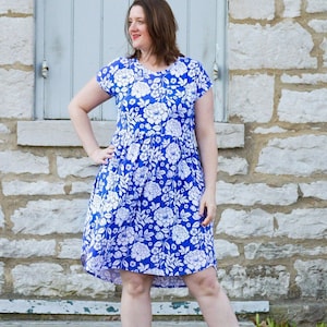 以下が含まれることがあります： 白い花柄の青いドレス。ドレスは半袖で丸いネックラインです。モデルは白いスニーカーを履いています。背景は石壁と閉じた窓です。