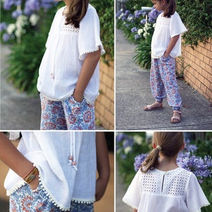 May include: A white cotton top with a crochet neckline and white pom-pom trim. The top has short sleeves and a relaxed fit. The model is wearing the top with blue and red floral print pants.