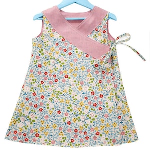 Könnte beinhalten: Ein ärmelloses Kleid mit floralem Muster, Wickeloberteil und Bindeband. Das Kleid hat ein hellblaues, gelbes, rotes und rosa Blumenmuster auf cremefarbenem Hintergrund. Das Oberteil ist einfarbig rosa und hängt an einem blauen Kleiderbügel.